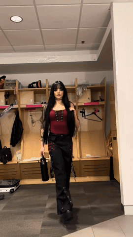A Woman In A Red Top And Black Pants Is Standing In A Locker Room Holding A Water Bottle GIF