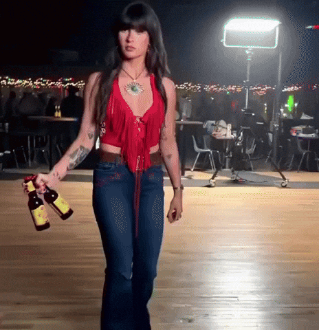 A Woman In A Red Top And Blue Jeans Holding Two Bottles Of Beer GIF
