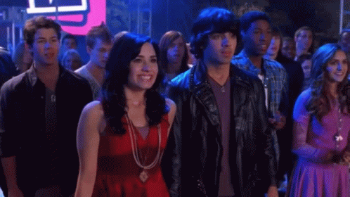 A Woman In A Red Top Stands Next To A Man In A Leather Jacket With Studs On It GIF