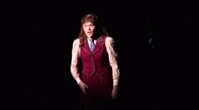 A Woman In A Red Vest And White Shirt Is Standing On A Stage In The Dark GIF