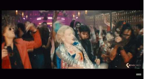 A Woman In A Sequined Jacket Is Dancing In Front Of A Crowd With The Word Kino On The Bottom Left GIF