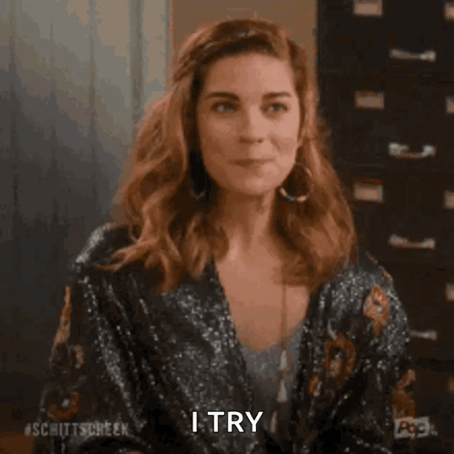 A Woman In A Sequined Jacket Is Smiling And Saying `` I Try '' While Holding Her Hair . GIF