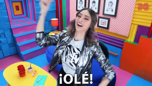 A Woman In A Silver Jacket Is Sitting In A Colorful Room With Her Arms In The Air And Says " Ole " GIF