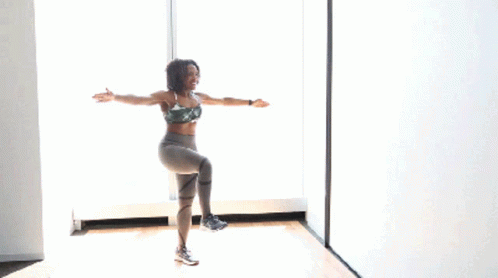 A Woman In A Sports Bra And Leggings Is Standing On One Leg In Front Of A Window With Her Arms In The Air GIF