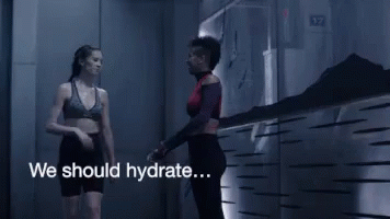 A Woman In A Sports Bra Is Walking Down A Hallway With The Words We Should Hydrate GIF