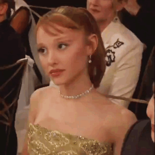 A Woman In A Strapless Gold Dress Is Sitting In A Crowd Of People . GIF