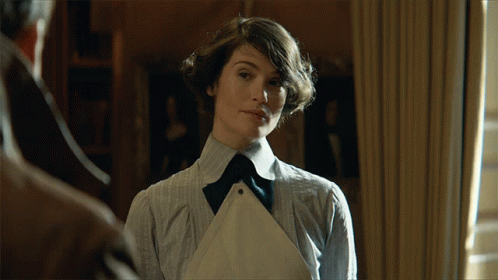 A Woman In A Striped Shirt And Apron Stands In Front Of A Window GIF
