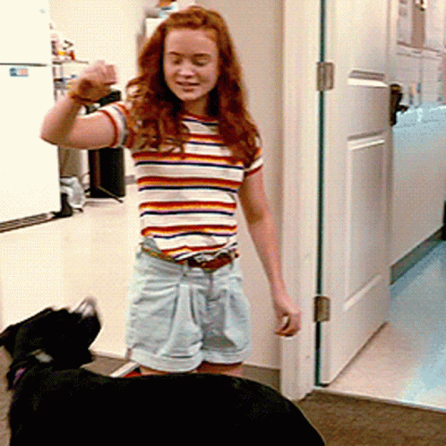 A Woman In A Striped Shirt And Shorts Stands Next To A Black Dog GIF