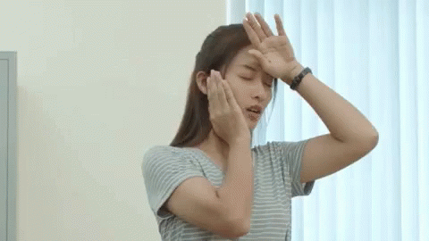 A Woman In A Striped Shirt Is Covering Her Face With Her Hands While Standing In Front Of A Window . GIF
