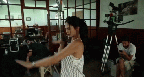 A Woman In A Striped Shirt Is Dancing In A Room With A Camera On A Tripod . GIF
