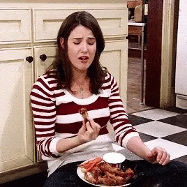 A Woman In A Striped Shirt Is Eating Chicken Wings In A Kitchen . GIF