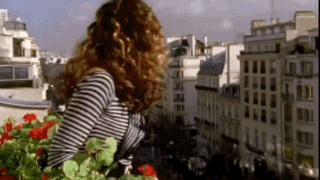 A Woman In A Striped Shirt Is Standing On A Balcony Looking Out Over A City GIF