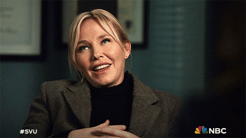 A Woman In A Suit And Black Turtleneck Is Smiling With The Nbc Logo Behind Her GIF