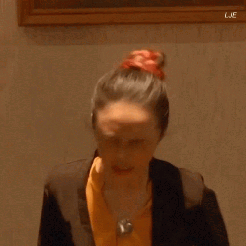 A Woman In A Suit And Orange Shirt Is Standing In Front Of A Framed Painting . GIF