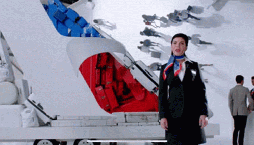 A Woman In A Suit And Scarf Stands In Front Of A Red And Blue Suitcase GIF