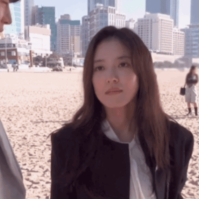 A Woman In A Suit And Tie Is Standing On A Sandy Beach GIF