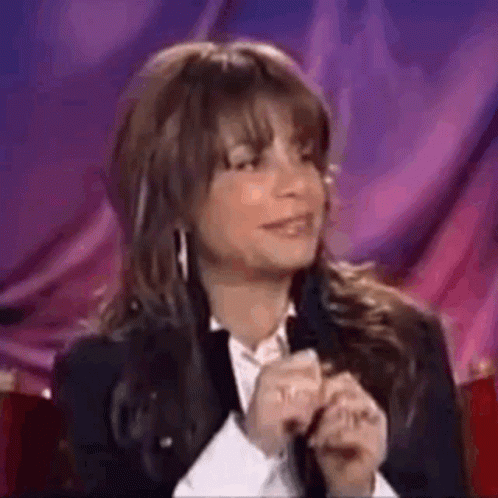 A Woman In A Suit And White Shirt Is Sitting In Front Of A Purple Curtain . GIF