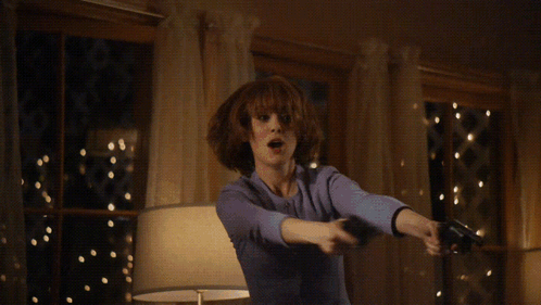 A Woman In A Suit Is Holding Two Guns In Front Of A Table GIF