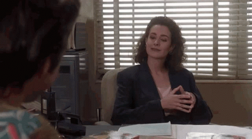 A Woman In A Suit Is Sitting At A Desk In Front Of A Window . GIF