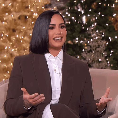 A Woman In A Suit Is Sitting In Front Of A Christmas Tree . GIF