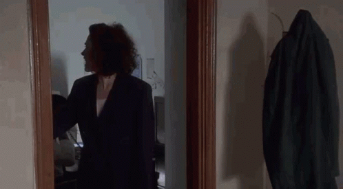 A Woman In A Suit Is Standing In A Hallway Next To A Coat Hanging On A Rack . GIF