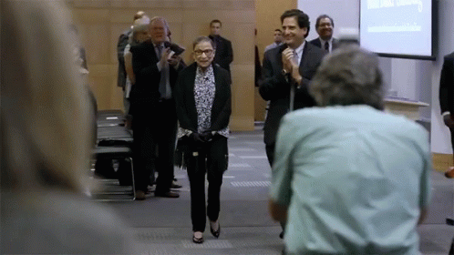 A Woman In A Suit Is Walking Down A Hallway With A Man Holding A Microphone Behind Her GIF
