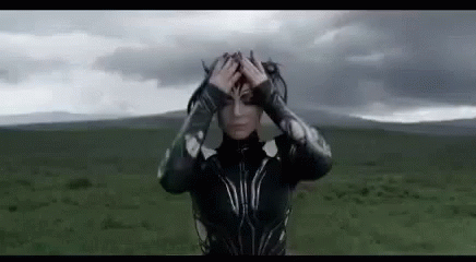 A Woman In A Superhero Costume Is Standing In A Field Holding Her Head . GIF