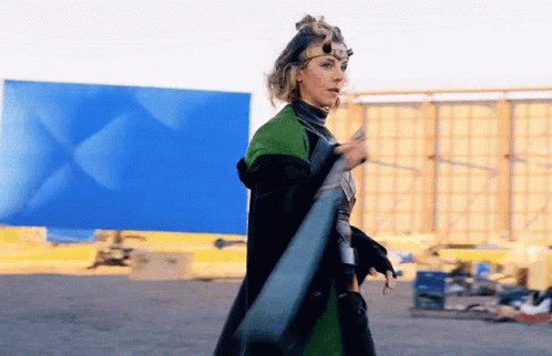 A Woman In A Superhero Costume Is Standing In Front Of A Blue Wall Holding A Sword . GIF