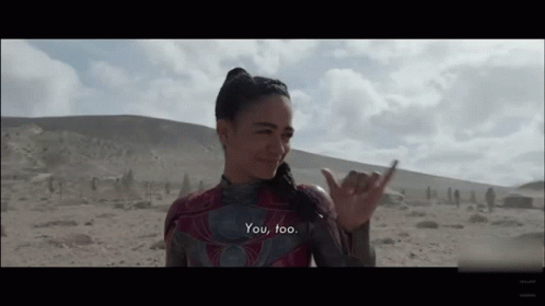 A Woman In A Superhero Costume Is Standing In The Desert And Making A Hawaiian Hand Sign . GIF