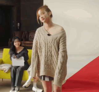 A Woman In A Sweater Is Standing In Front Of A Woman Sitting On A Yellow Couch GIF
