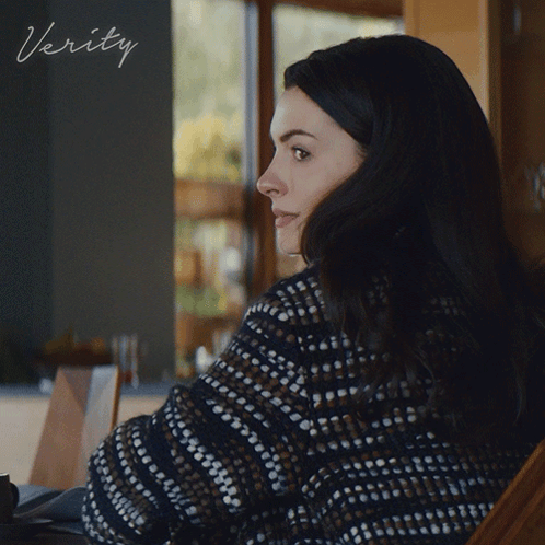 A Woman In A Sweater Sits In Front Of A Sign That Says ' Verity ' On It GIF