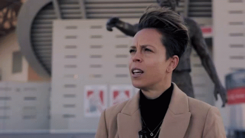 A Woman In A Tan Coat Stands In Front Of A Statue Of A Soccer Player GIF
