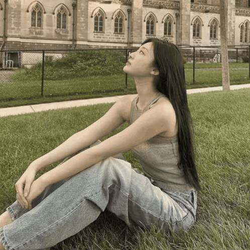 A Woman In A Tank Top And Jeans Is Sitting In The Grass GIF