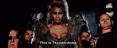 A Woman In A Thunderdome Costume Is Surrounded By Men GIF