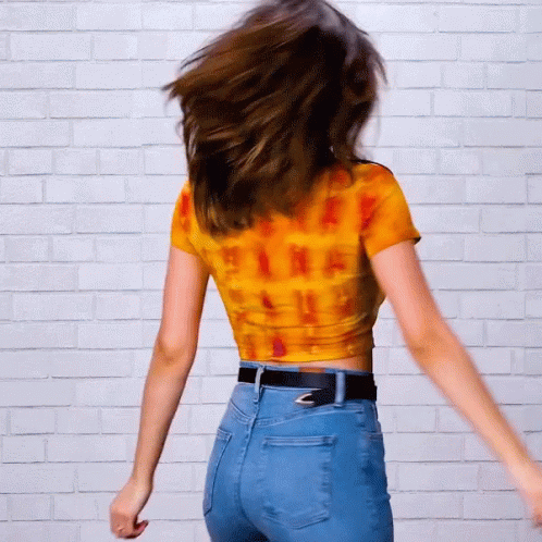 A Woman In A Tie Dye Shirt And Blue Jeans Is Dancing In Front Of A White Brick Wall GIF