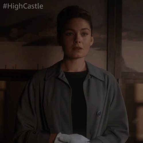 A Woman In A Trench Coat And White Gloves Stands In Front Of A Sign That Says High Castle GIF