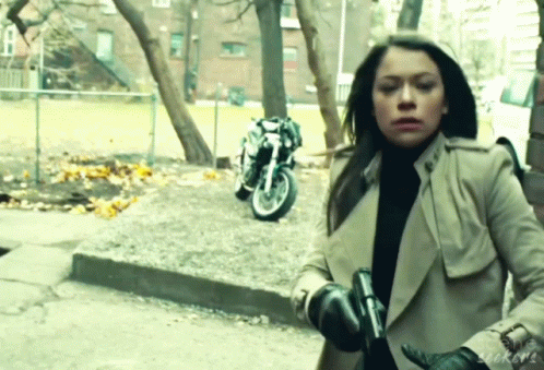 a woman in a trench coat holds a gun in front of a parked motorcycle