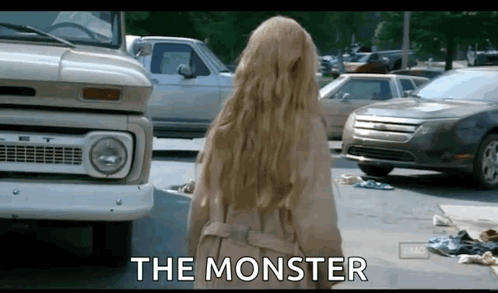 A Woman In A Trench Coat Is Standing In Front Of A Truck And Cars And The Words The Monster Are Written On The Screen . GIF