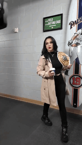 A Woman In A Trench Coat Stands In Front Of A Sign That Says " Field To The Batting Cage " GIF