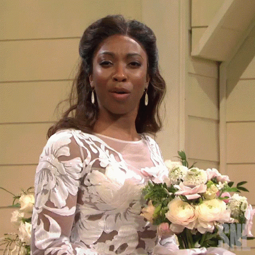 A Woman In A Wedding Dress Holds A Bouquet Of Flowers In Front Of A Sign That Says Nbc GIF