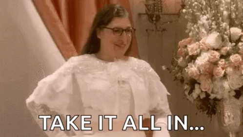 A Woman In A Wedding Dress Is Standing In Front Of A Vase Of Flowers And Says `` Take It All In '' . GIF