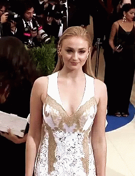 A Woman In A White And Gold Dress Stands In Front Of Photographers GIF