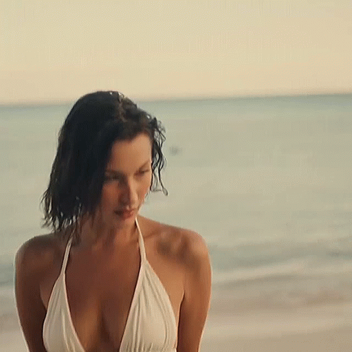 A Woman In A White Bikini Is Standing On The Beach Near The Ocean . GIF