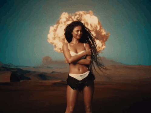 A Woman In A White Bra And Black Shorts Is Standing In Front Of An Explosion GIF