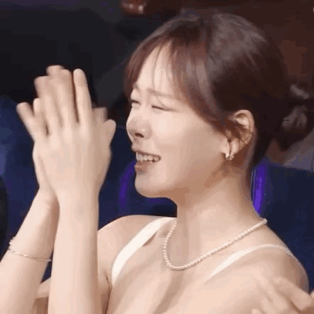 A Woman In A White Bra And Pearl Necklace Is Clapping Her Hands In A Theater . GIF