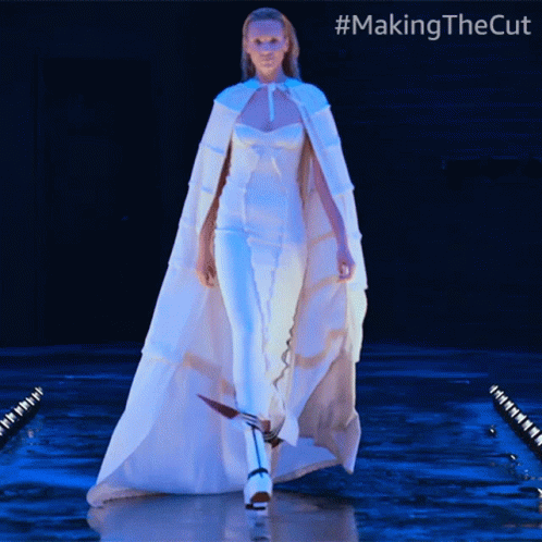 A Woman In A White Cape Is Walking Down A Runway With #Makingthecut Written On The Bottom GIF