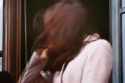 A Woman In A White Coat Is Standing In Front Of A Window Holding Her Hair . GIF