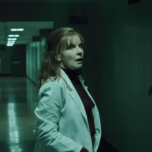 A Woman In A White Coat Stands In A Hallway GIF