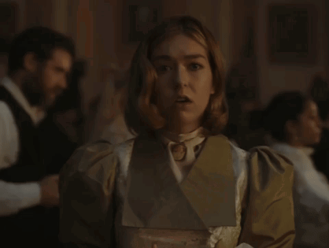 A Woman In A White Dress And A Brown Jacket Stands In A Dark Room GIF