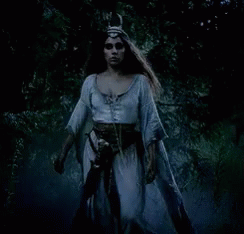 A Woman In A White Dress And A Horned Hat Is Walking Through A Dark Forest . GIF